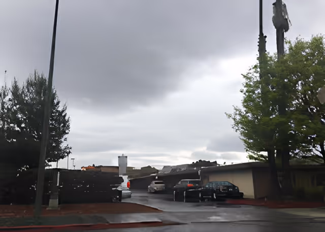 Outdoor view of a parking area at a facility with several parked cars, trees on both sides, and a cloudy sky overhead.