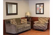 A cozy sitting area with a brown sofa and a patterned armchair, a wooden side table with a lamp, a framed mirror on the wall above the sofa, and a framed abstract painting on the adjacent wall.