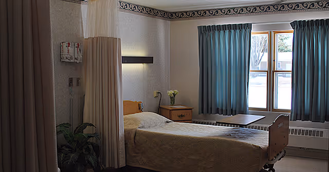 A simple, clean hospital-style bedroom with a single bed covered in a beige quilt, a wooden nightstand with a small vase of flowers, a window with blue curtains, and a privacy curtain partially drawn around the bed.