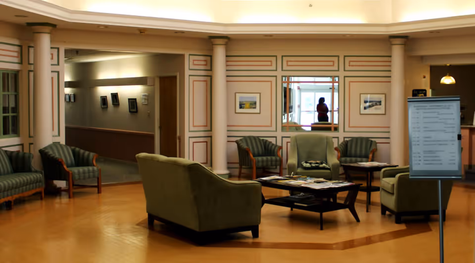 A spacious and well-lit common area with several green upholstered chairs and sofas arranged around a central coffee table holding magazines. The room features decorative columns and framed artwork on the walls. A hallway is visible in the background, and a person is reflected in a mirror on the far wall. There is also a signboard with a schedule or information near the seating area.