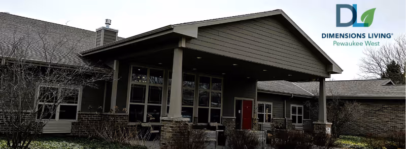 Exterior view of a single-story building with a covered entrance supported by columns. The building has large windows and a combination of brick and siding on the walls. Leafless shrubs and trees are visible in the foreground under an overcast sky.