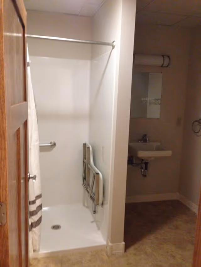 A small bathroom with a walk-in shower featuring a grab bar and a foldable shower seat mounted on the wall. To the right of the shower is a small sink with a mirror above it and a light fixture. The floor has a beige tile pattern, and the walls are painted white.