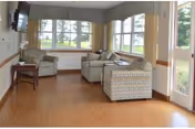A bright living room area with large windows letting in natural light. The room contains three cushioned armchairs arranged around a small wooden table. A flat-screen TV is mounted on the wall to the left, and the floor is covered with wood-like flooring. Outside greenery is visible through the windows.