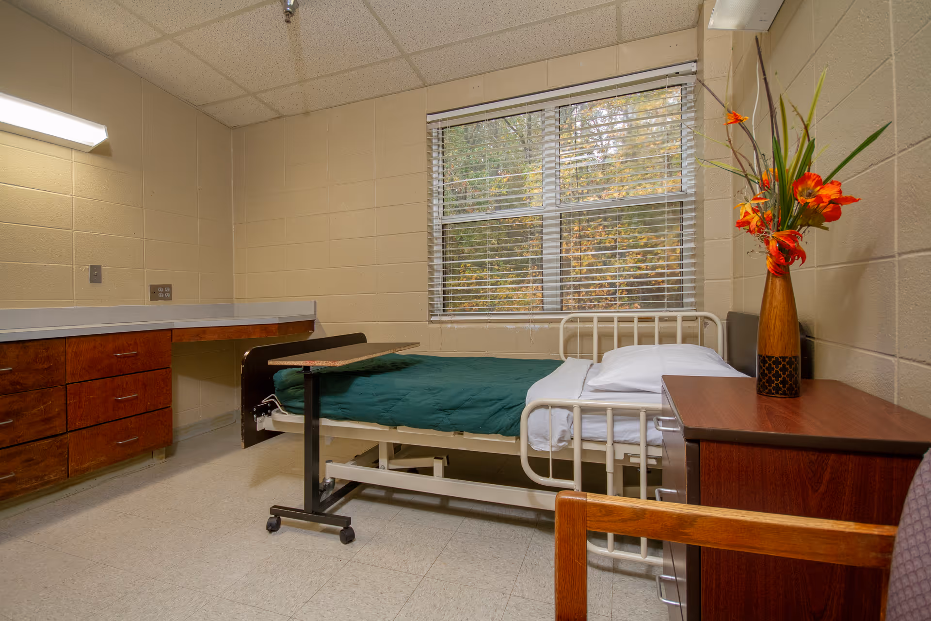 A simple assisted-living bedroom with a single hospital-style bed, overbed table, dresser, window with blinds, and a vase of flowers on a nightstand.