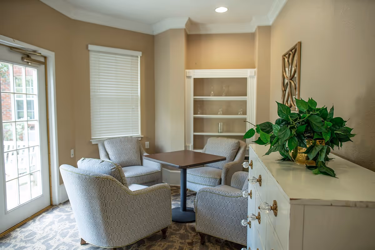 A cozy sitting area with four patterned armchairs arranged around a square wooden table. The room has beige walls, a window with closed blinds, a glass door leading outside, a white cabinet with decorative knobs and a green potted plant on top, and a white built-in shelf with glassware.