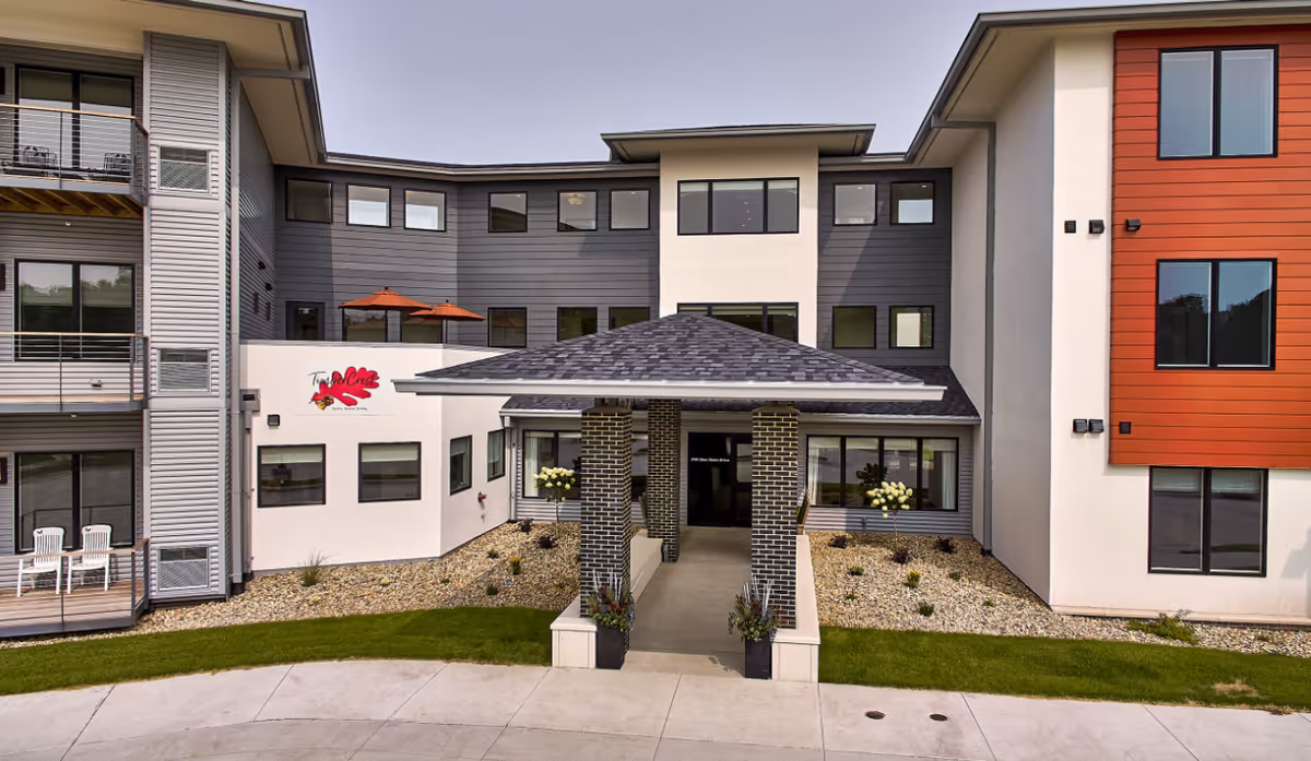 Front exterior view of TimberCrest at Glen Oaks, a modern multi-story senior living facility with a covered entrance supported by brick pillars, landscaped areas with rocks and plants, and balconies with chairs and umbrellas.