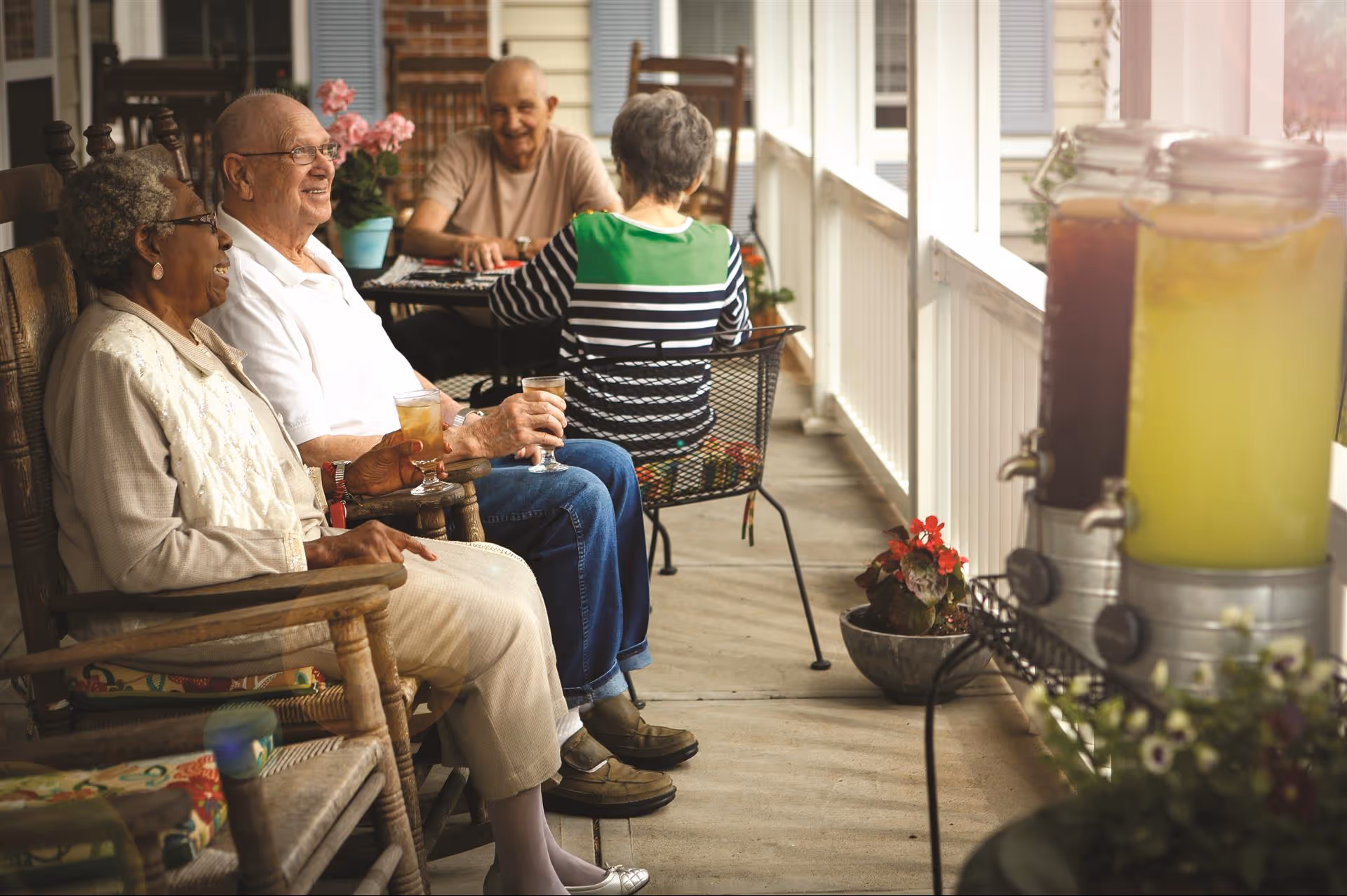 Four elderly people sitting and enjoying drinks on a covered porch. Two are seated in rocking chairs in the foreground, smiling and holding glasses with beverages. Two others are seated at a table in the background, engaged in conversation. The porch has potted plants and beverage dispensers with lemonade and iced tea on a stand to the right.
