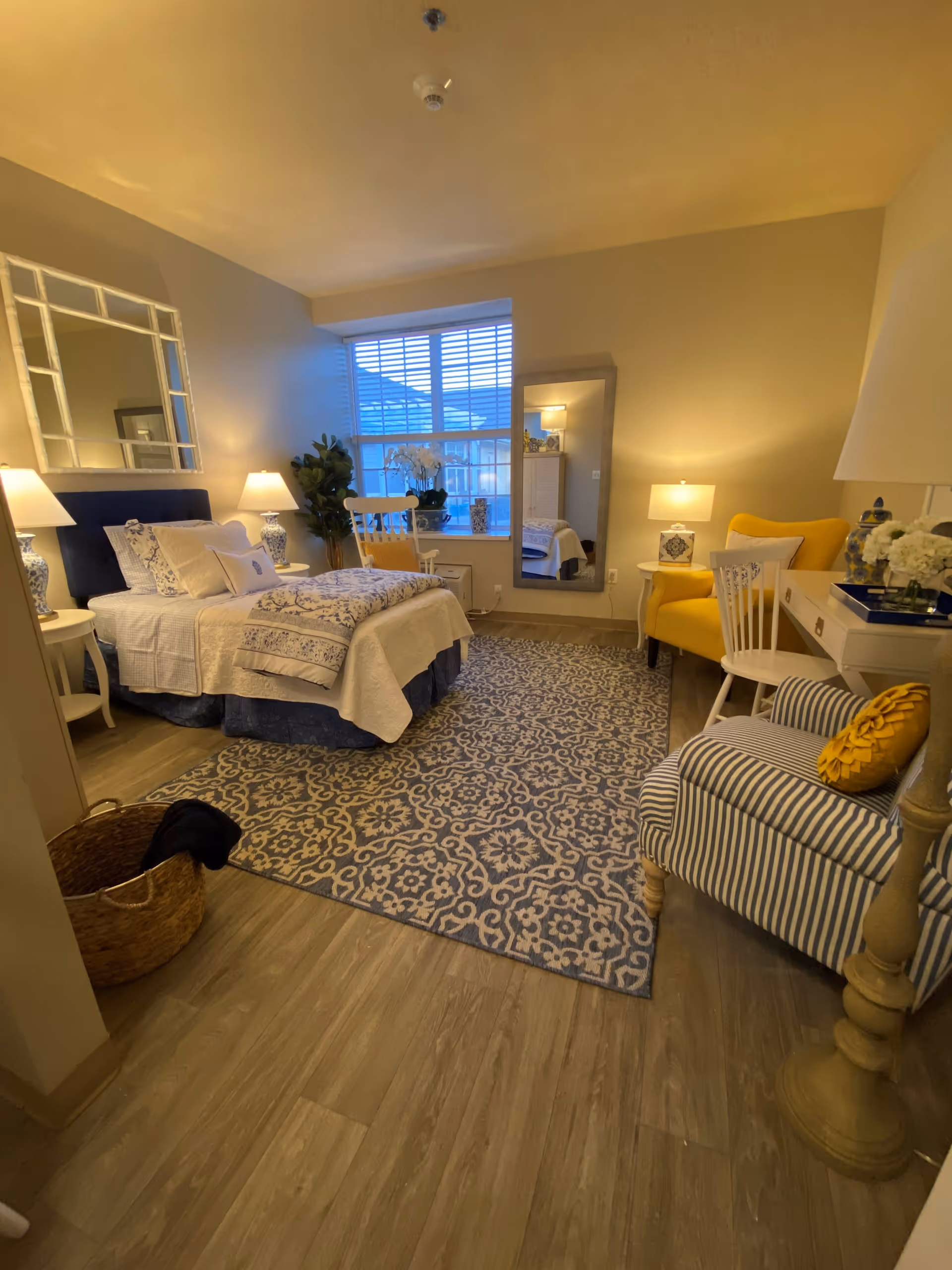 A cozy bedroom with a bed dressed in white and blue bedding, two bedside tables with lamps, a large patterned rug, a striped armchair with a yellow pillow, a yellow armchair, a white desk with a chair, a tall floor lamp, a large mirror on the wall, and a window with blinds letting in natural light.