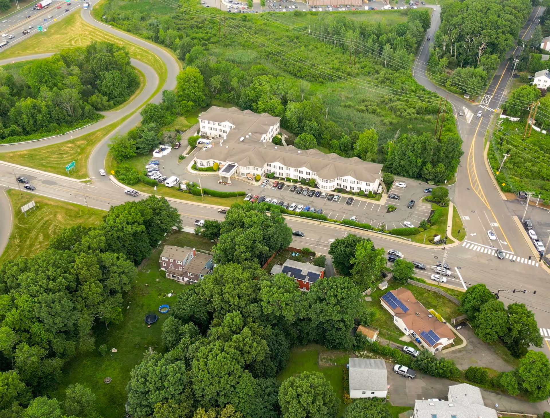 Aerial view of Benchmark Senior Living at Waltham Crossings facility surrounded by trees and greenery, with a parking lot and nearby roads including a curved highway and intersections.