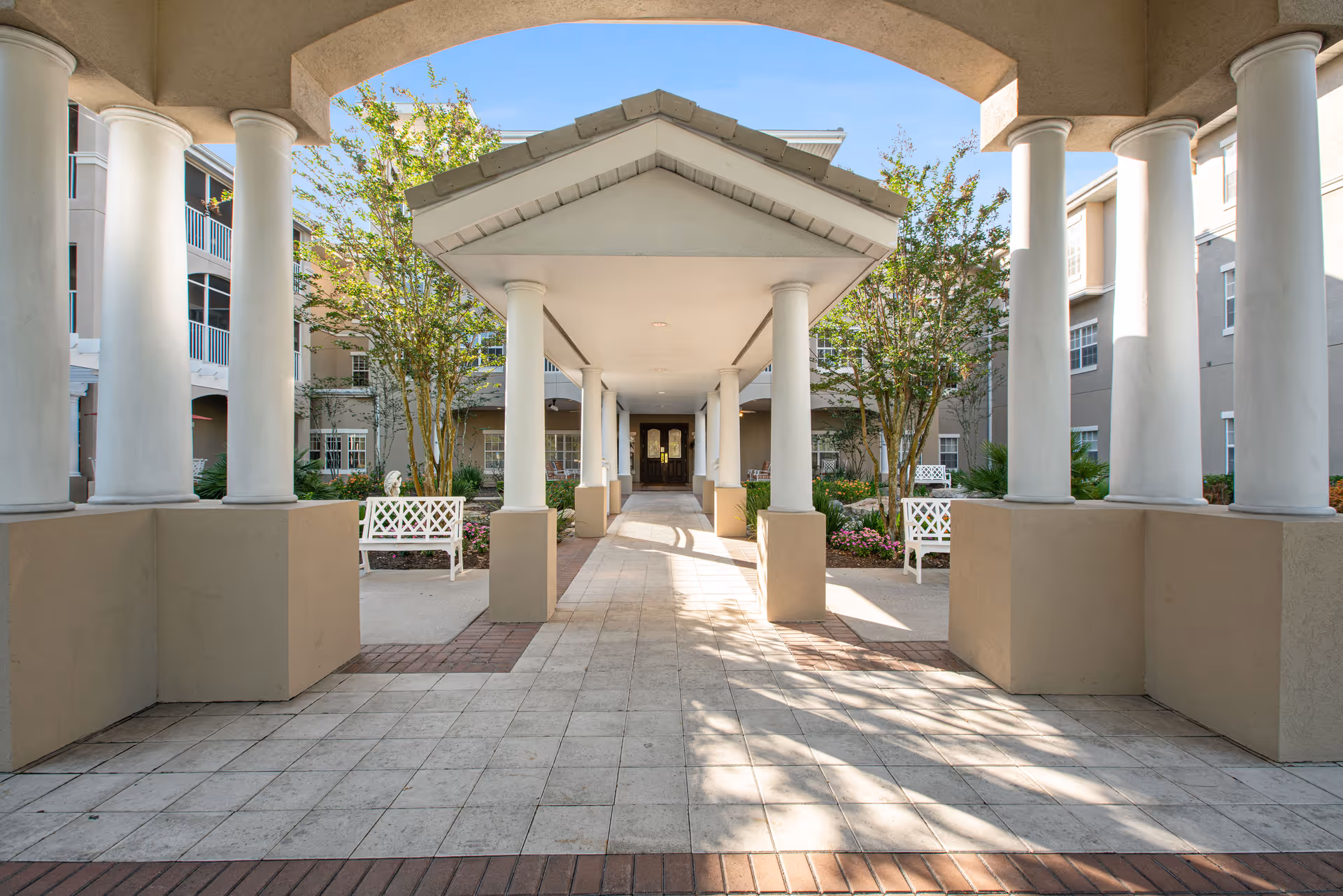 Covered entrance walkway with white columns, benches, and landscaping leading to the front doors of a multi-story residence.