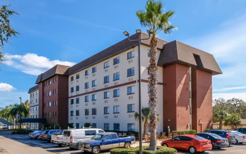 Exterior view of a multi-story senior living facility building with a parking lot in front. Several cars are parked, and palm trees are visible near the building under a clear blue sky.