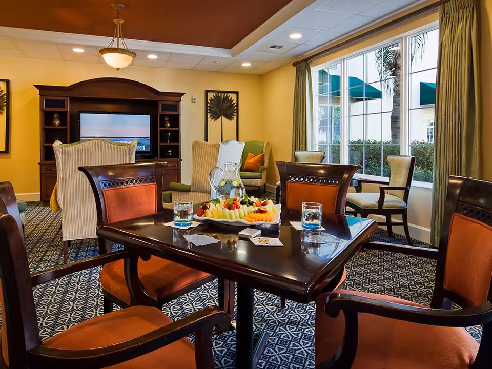 Communal dining and lounge area with a polished wooden table topped with water glasses and a fruit platter, upholstered chairs, armchairs, a TV cabinet, and large windows.