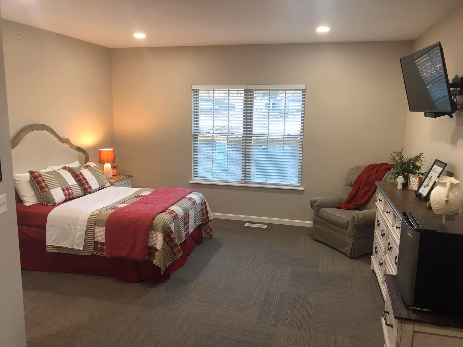 A cozy bedroom with a bed, nightstand, window, chair, dresser, and TV.