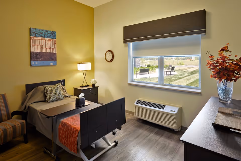 A well-lit senior living bedroom with a single bed featuring a brown headboard and footboard, a beige blanket, and a decorative pillow. Next to the bed is a small black nightstand with a lamp and a small flower arrangement. A striped armchair is positioned to the left of the bed. A window with a dark valance shows an outdoor patio area with chairs and greenery. On the right side, there is a dark wooden dresser with a vase of orange flowers and a decorative tray. The room has light yellow walls and wood flooring.