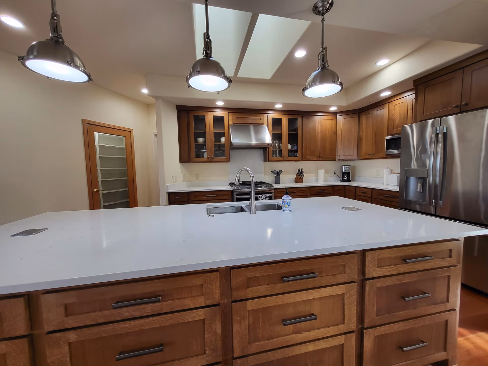 Modern kitchen with wooden cabinets, a large white island countertop with a built-in sink and faucet, stainless steel refrigerator, stove with a hood, microwave, and three hanging pendant lights above the island. There is a door with frosted glass panels on the left side and recessed ceiling lights.