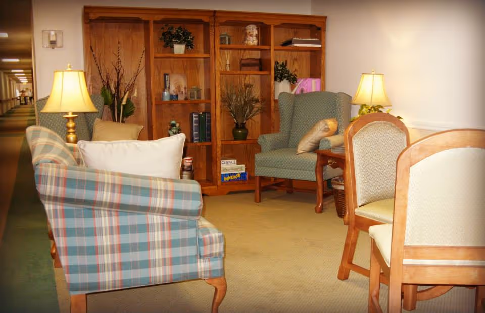 A cozy sitting area in a senior living facility featuring a plaid upholstered armchair with a white pillow, a green wingback chair with a beige pillow, two wooden side tables with lamps, a wooden bookshelf with decorative items and books, and a wooden dining table with upholstered chairs. The area is carpeted and located near a hallway.