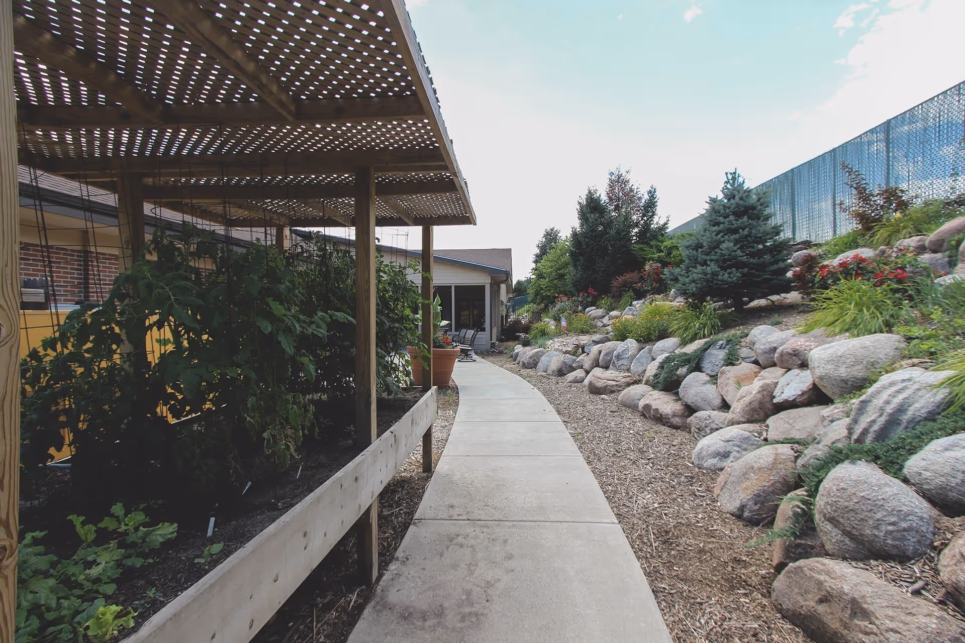 A paved walkway runs between a raised garden bed with plants under a wooden lattice structure on the left and a landscaped rock garden with shrubs and flowers on the right, leading to a building in the background under a partly cloudy sky.