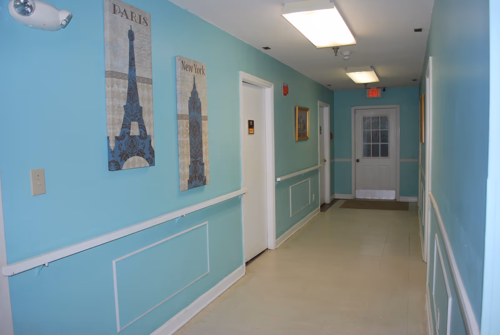 A clean, well-lit hallway with light blue walls and white trim. The hallway has several closed white doors, handrails along the walls, and framed artwork featuring the Eiffel Tower labeled 'Paris' and a New York skyscraper labeled 'New York'. There is an exit sign above a door at the end of the hallway.