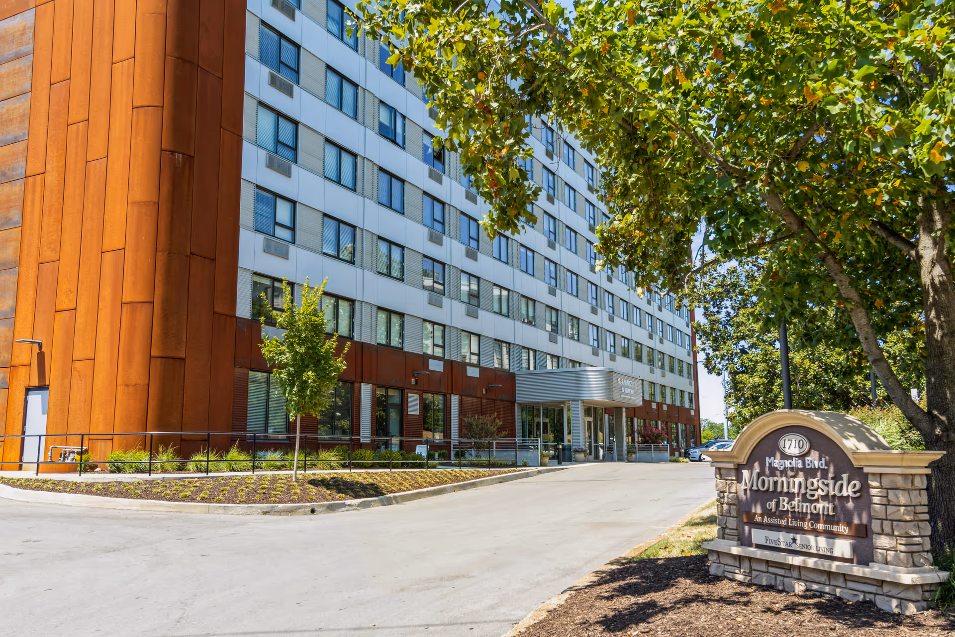 Exterior view of Morningside of Belmont, an assisted living community, showing a multi-story building with a modern facade and a sign at the entrance surrounded by trees and landscaping.