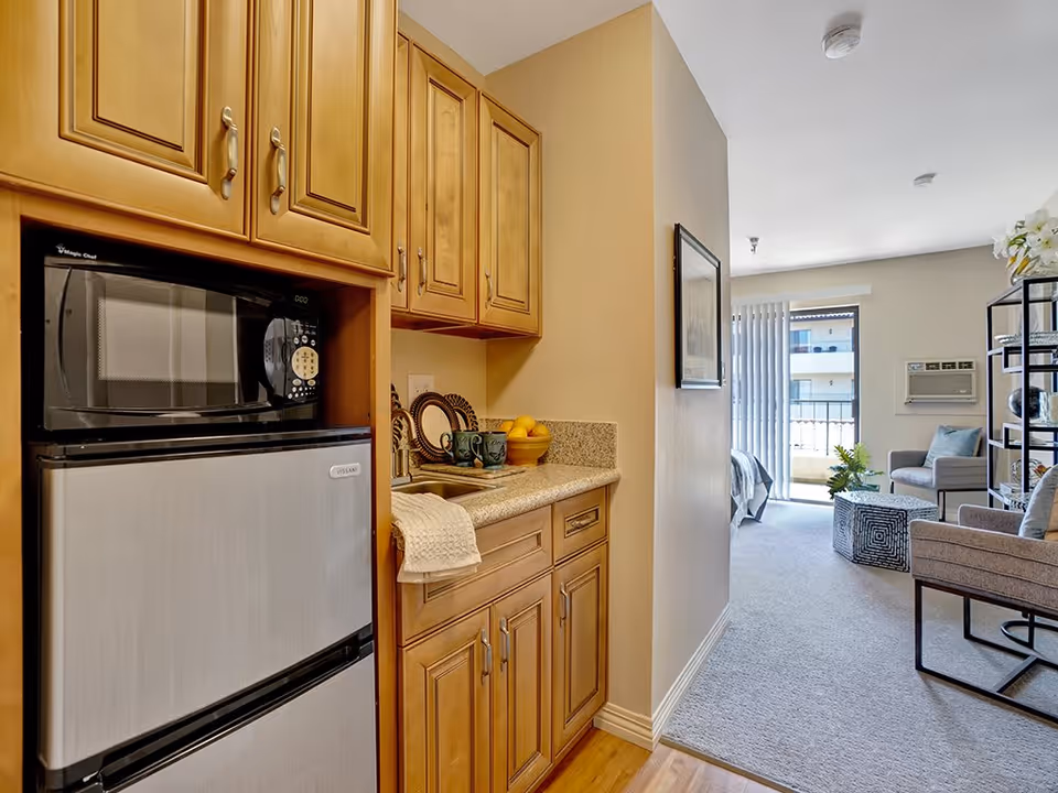View of a small kitchen area with wooden cabinets, a microwave, and a mini refrigerator on the left. The kitchen counter has a sink, two mugs, a bowl of fruit, and a towel hanging from the cabinet. Beyond the kitchen, there is a living area with carpeted flooring, a chair, a small table, a shelving unit, and a sliding glass door leading to a balcony.