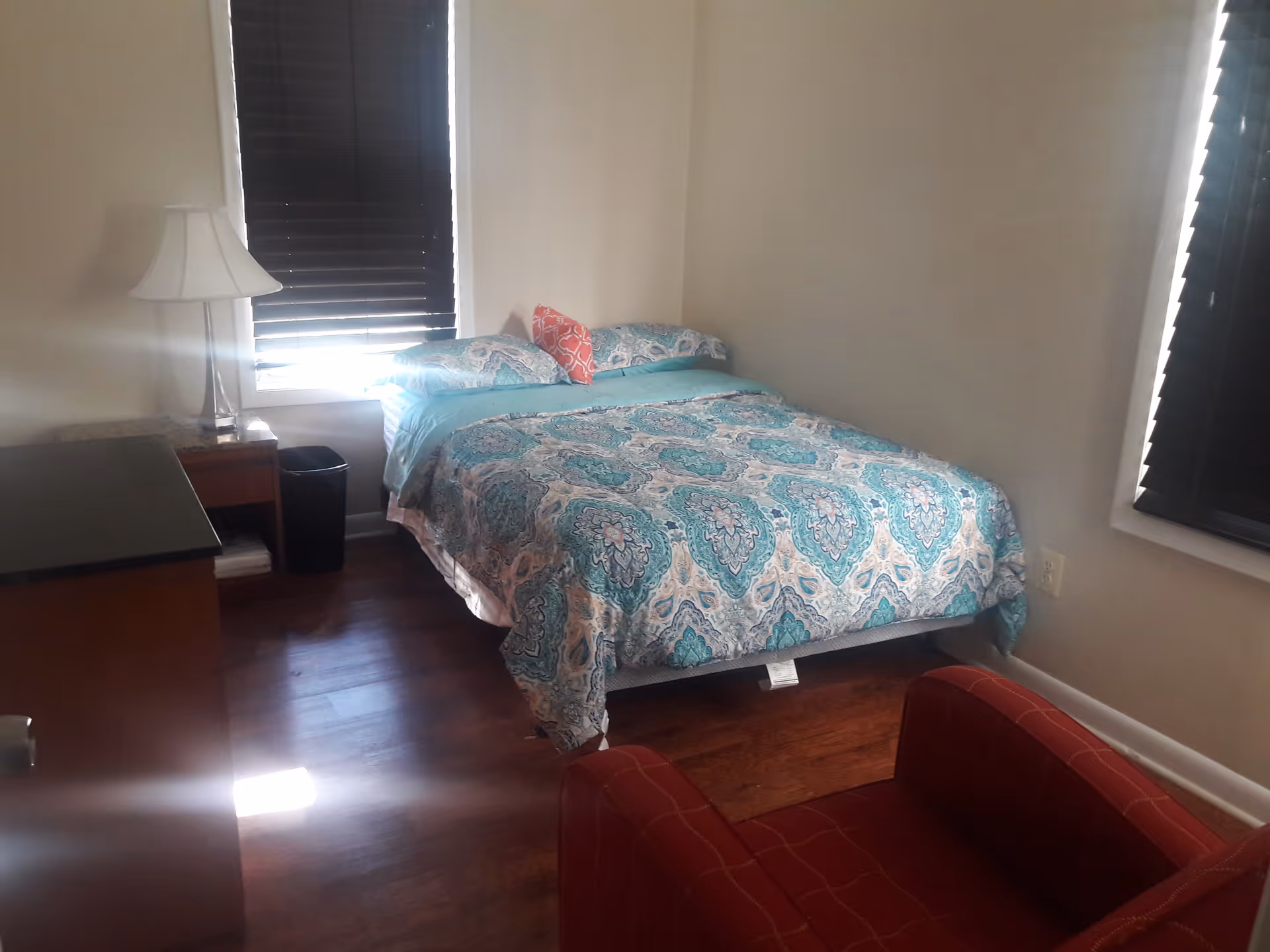 A small bedroom with a bed covered in a blue and white patterned comforter and pillows, a red armchair in the foreground, a wooden dresser with a lamp on top, and two windows with dark blinds.
