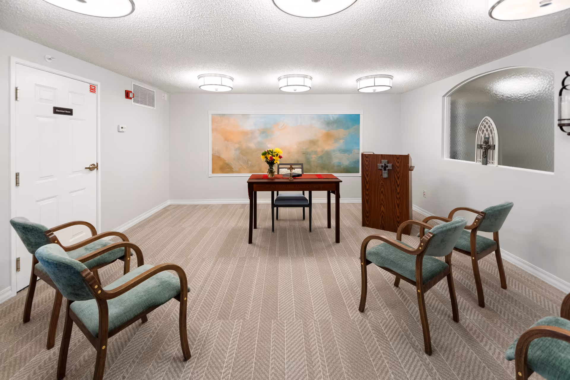 A small meeting or chapel room with six green upholstered chairs arranged in two rows facing a wooden table with a vase of flowers and a chair behind it. There is a wooden podium with a cross on the front to the right of the table. The room has beige carpet, white walls, three ceiling lights, a large abstract painting on the back wall, and a frosted glass window with a cross design on the right wall.