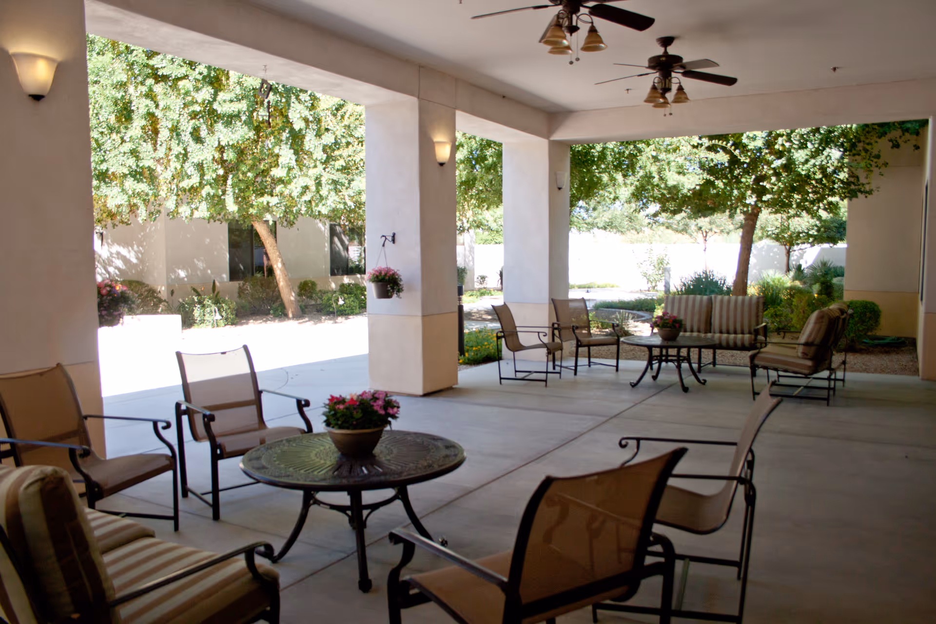 Covered outdoor patio area with several metal-framed chairs and tables, some with cushions and flower pots on top. The patio overlooks a garden with trees and shrubs, and ceiling fans are mounted on the patio ceiling.