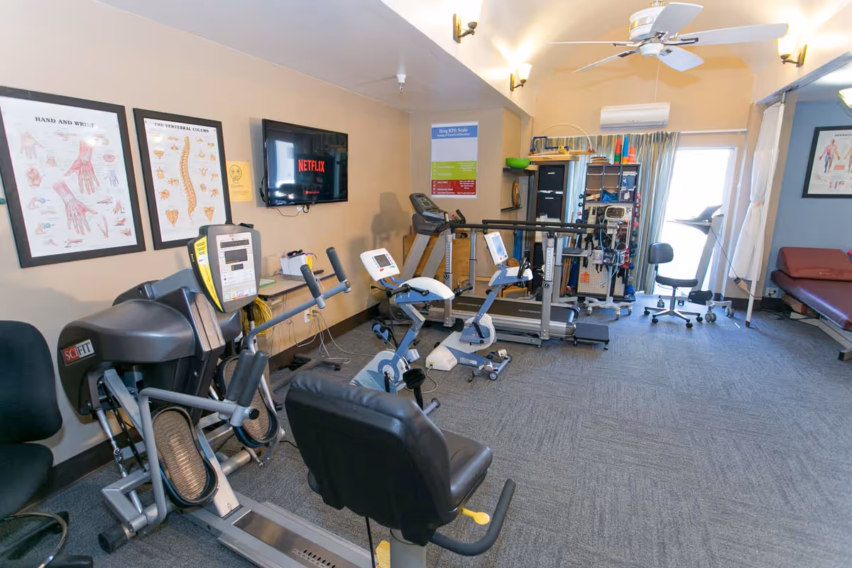 A rehabilitation or physical therapy room with exercise equipment including stationary bikes, a treadmill, and other fitness machines. The room has anatomical charts on the wall, a wall-mounted TV displaying Netflix, a ceiling fan, and various therapy tools and equipment. There is a window with curtains and a treatment table in the corner.