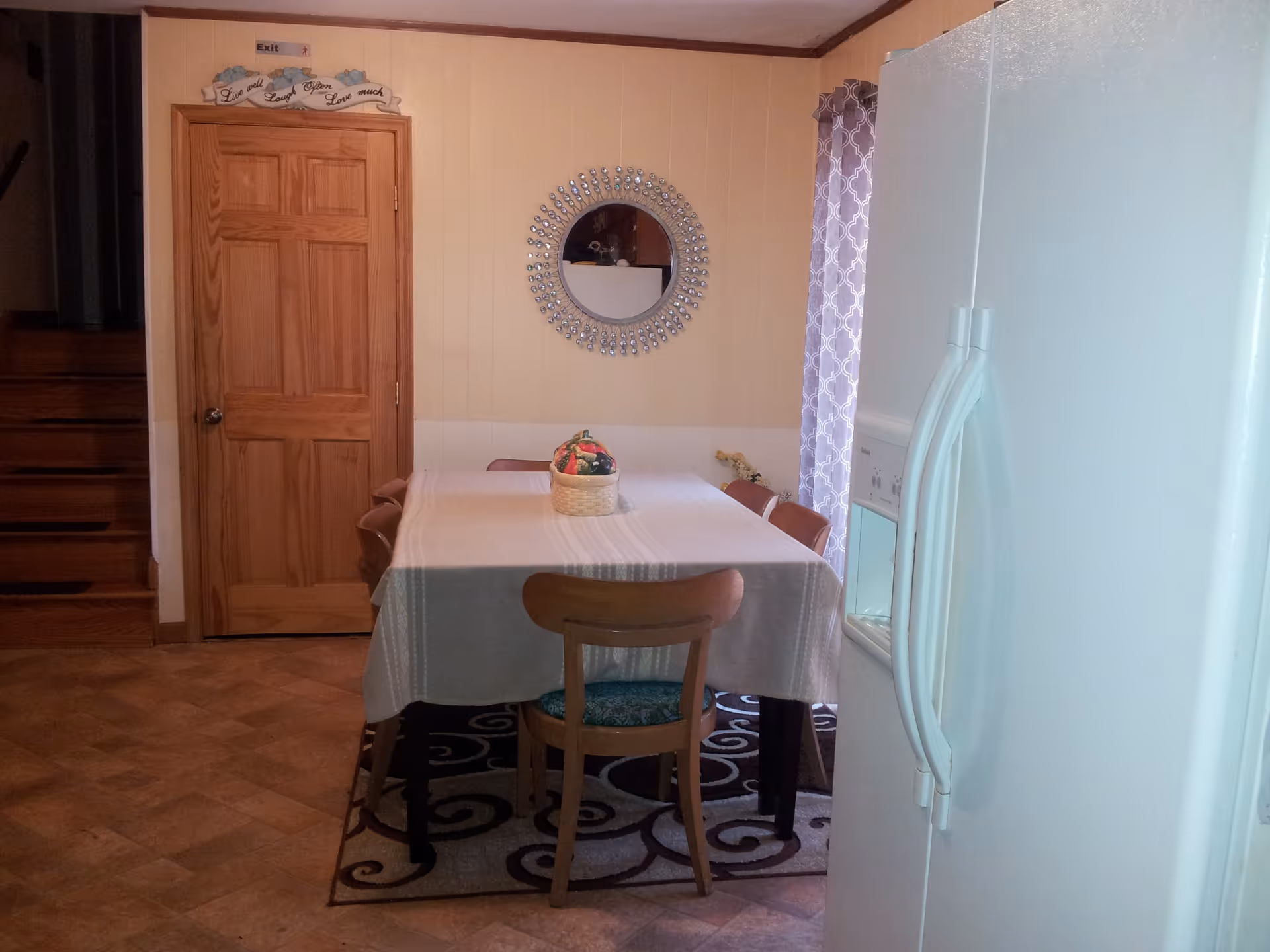 A dining area with a rectangular table covered with a white tablecloth and surrounded by wooden chairs. A decorative basket sits in the center of the table. Behind the table, there is a wooden door with a sign above it that reads 'Exit' and a decorative plaque with the words 'Live well, Laugh often, Love much.' A round mirror with a decorative frame hangs on the wall. To the right, a white refrigerator is partially visible, and a window with patterned curtains lets in natural light.
