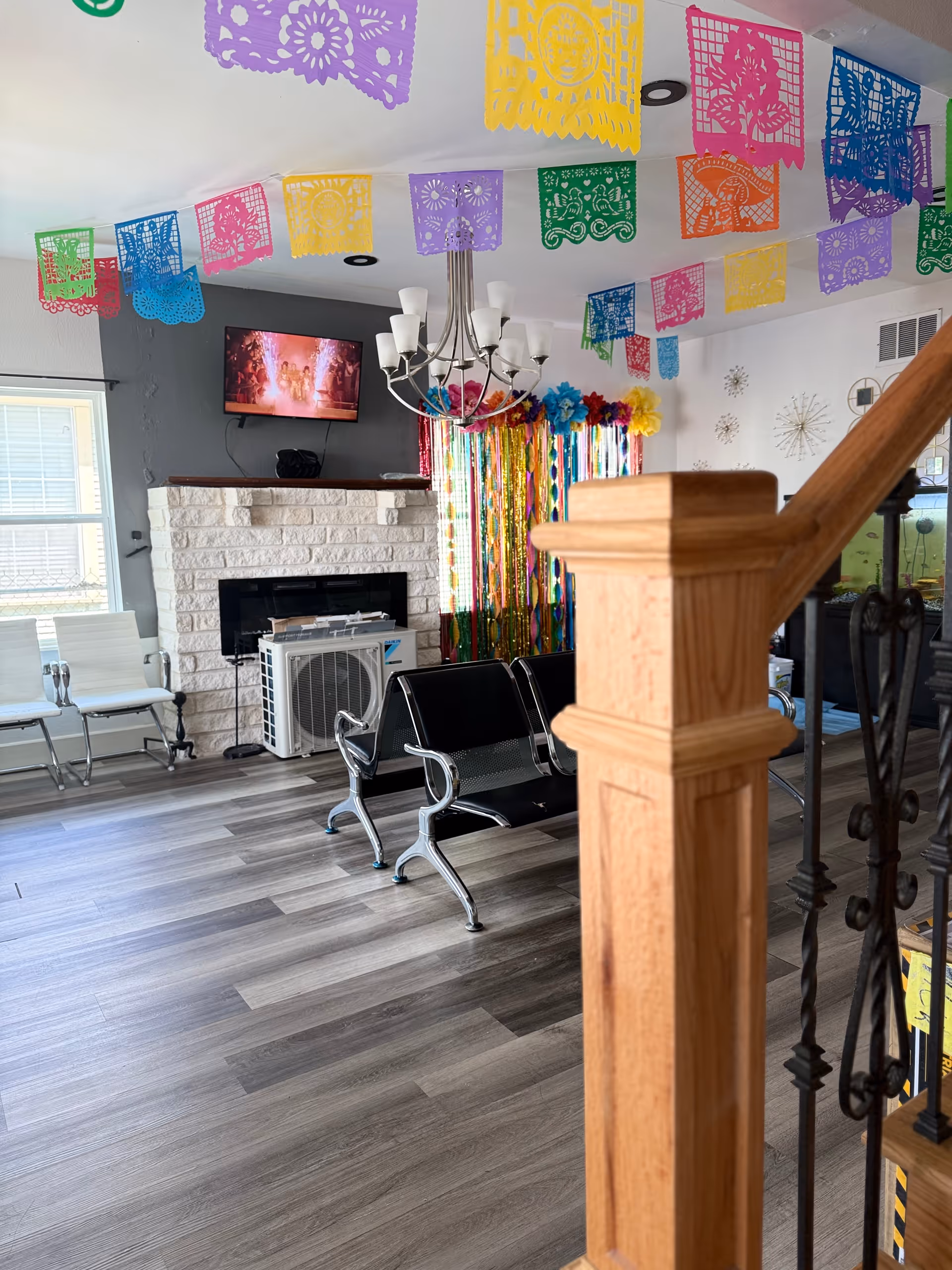 A decorated common room with colorful hanging paper banners, metal waiting chairs, a fireplace with a TV above it, and a wooden stair post in the foreground.