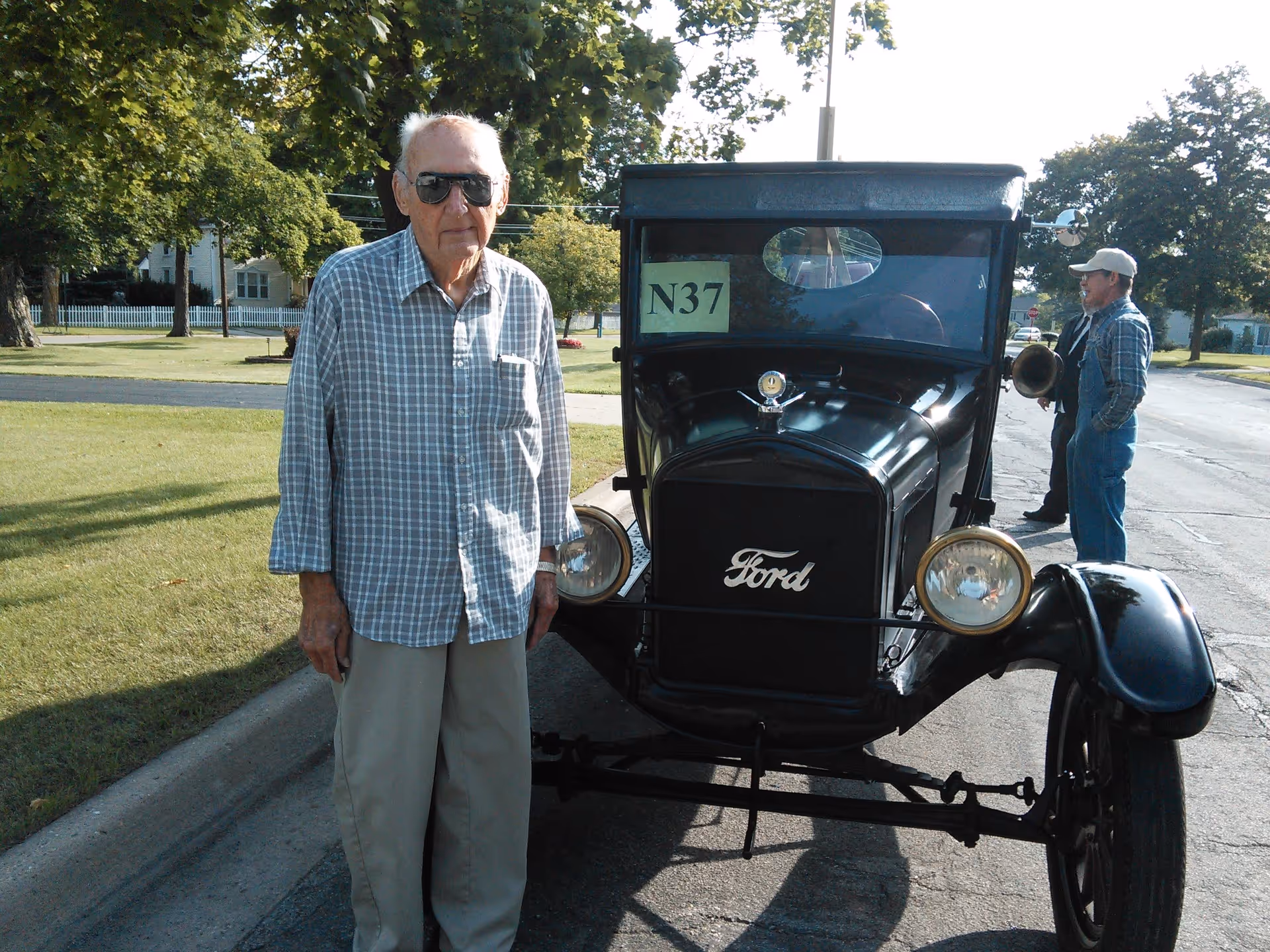 An elderly man wearing sunglasses and a checkered shirt stands next to a vintage black Ford car with a sign labeled N37 on the windshield. In the background, two men are talking on a street lined with trees and houses.