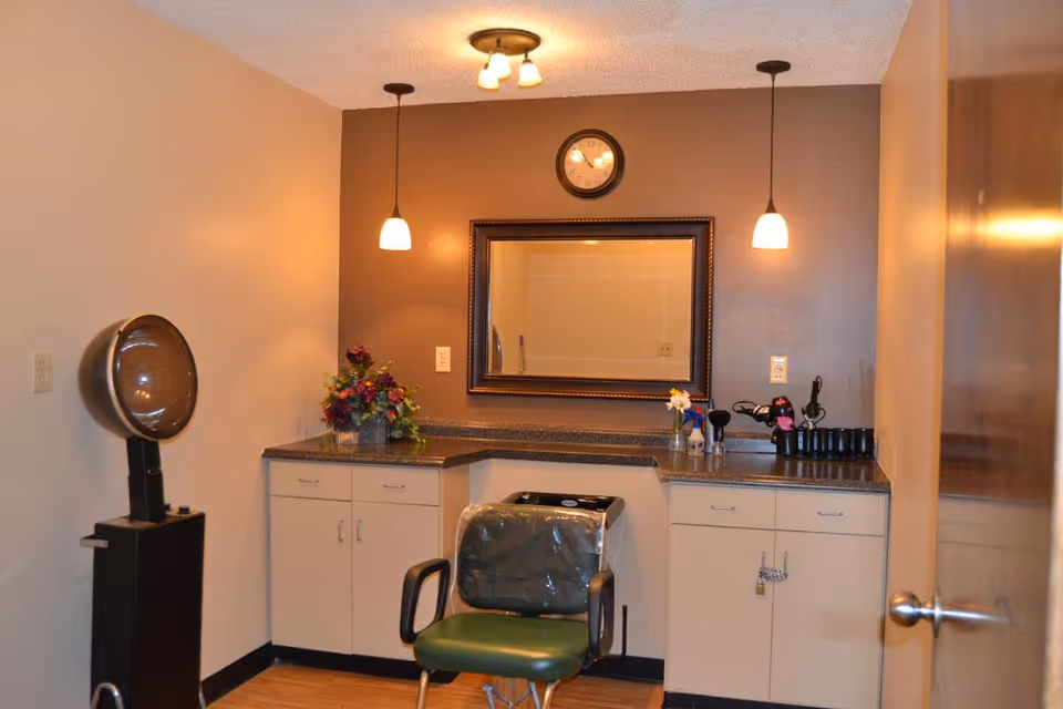 Small salon-style room with a hooded hair dryer, a plastic-covered styling chair, countertop and cabinets under a mirror and pendant lights.