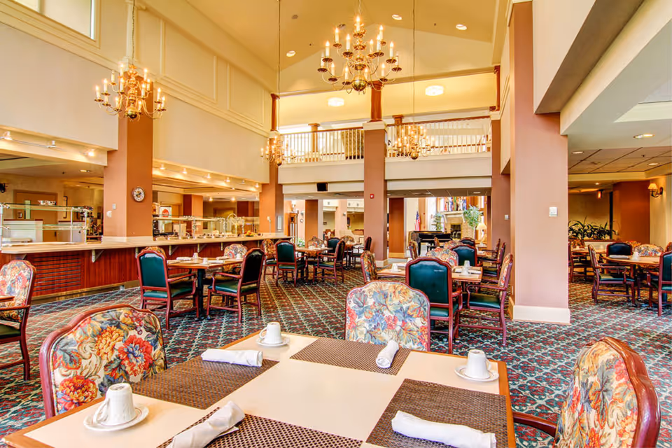 A spacious dining room with high ceilings and multiple chandeliers. The room features tables set with placemats, cups, and rolled napkins. Chairs have floral upholstery and dark wood frames. The carpet has a patterned design, and there is a buffet area along one wall. The room has a warm, inviting atmosphere with beige walls and large columns.