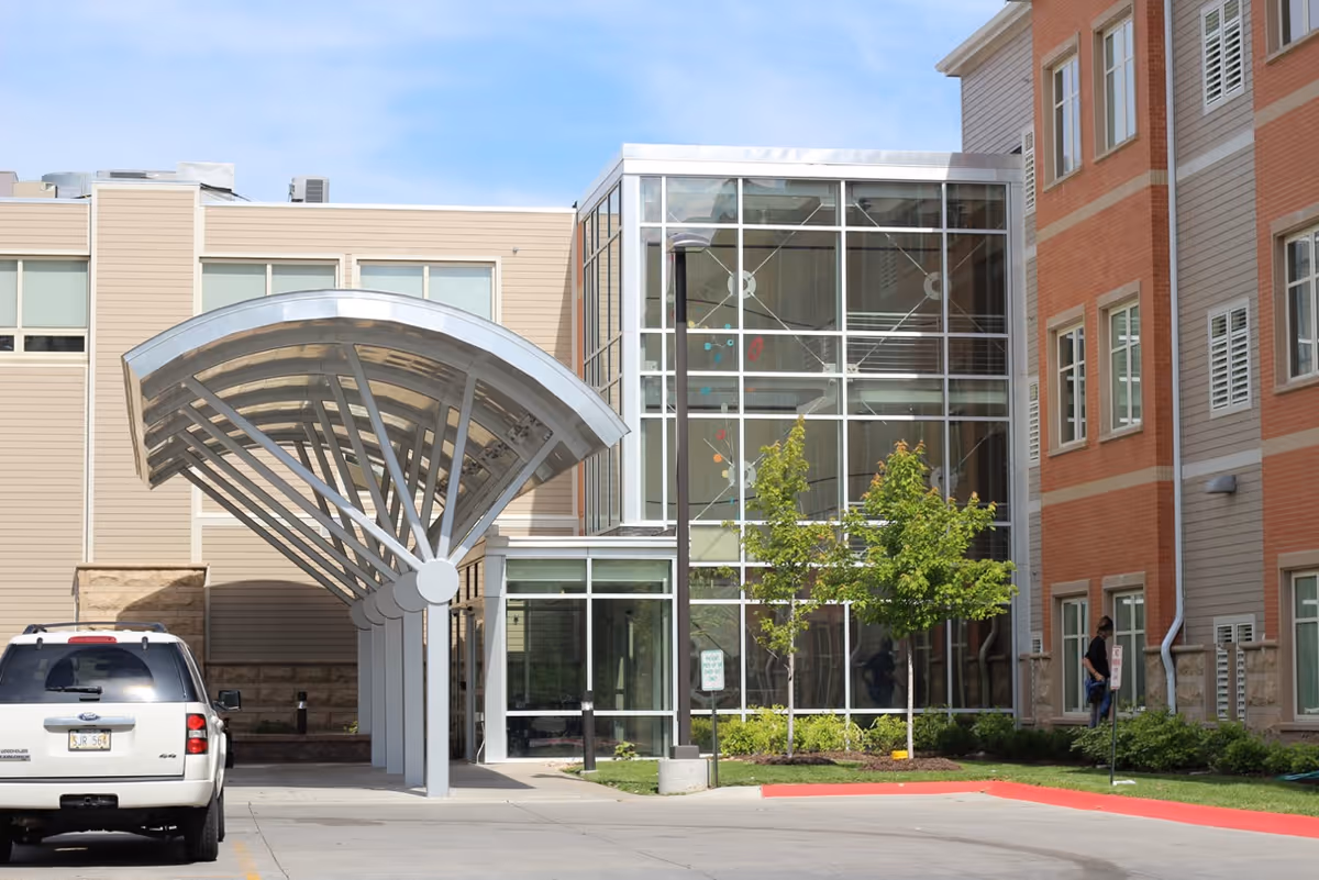 Entrance of a senior living building featuring a metal covered drop-off canopy, glass lobby, parked SUV, and brick and siding façade.