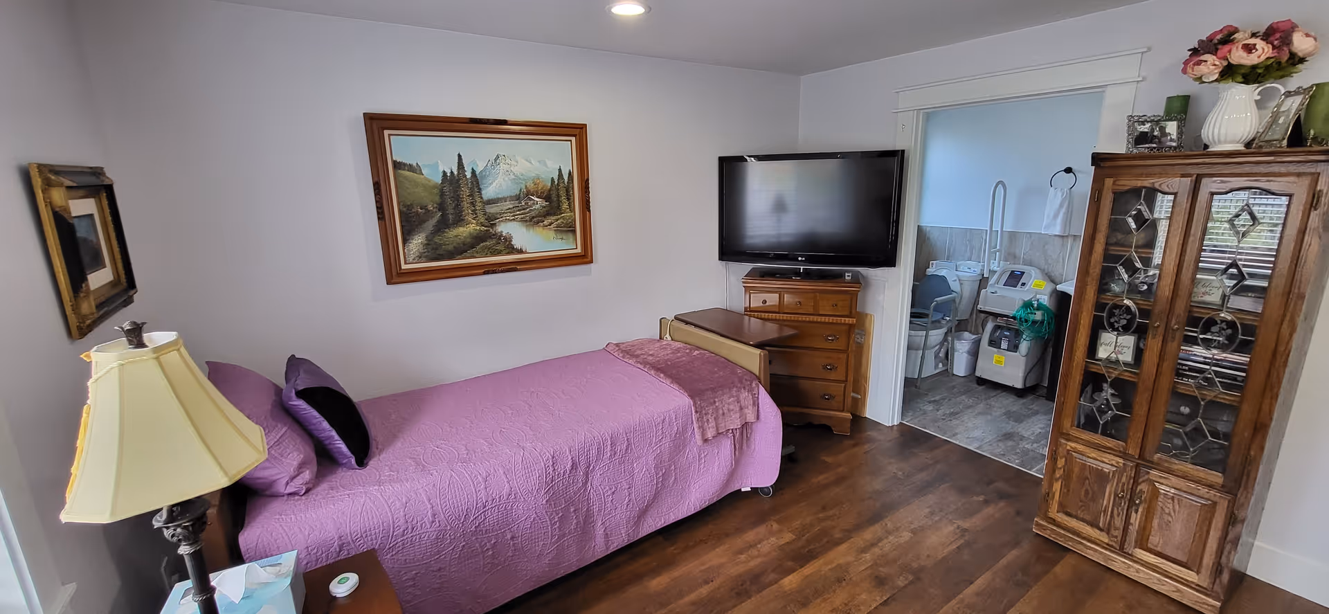 A bedroom with a single bed covered in a purple quilt and matching pillows. There is a wooden nightstand with a lamp and tissues next to the bed. A framed landscape painting hangs on the wall above the bed. Across from the bed is a wooden dresser with a flat-screen TV mounted above it. To the right, there is a wooden cabinet with glass doors displaying decorative items. An adjoining room with medical equipment is visible through an open doorway. The floor is wooden and the walls are painted white.