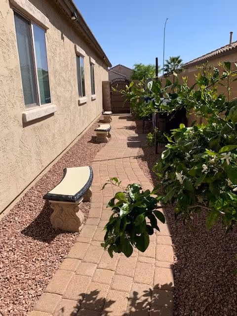 A narrow outdoor pathway with a curved brick walkway bordered by red gravel. There are two stone benches along the left side next to a beige stucco building with windows. On the right side, there is a tall fence and green leafy plants with white flowers. The sky is clear and blue.