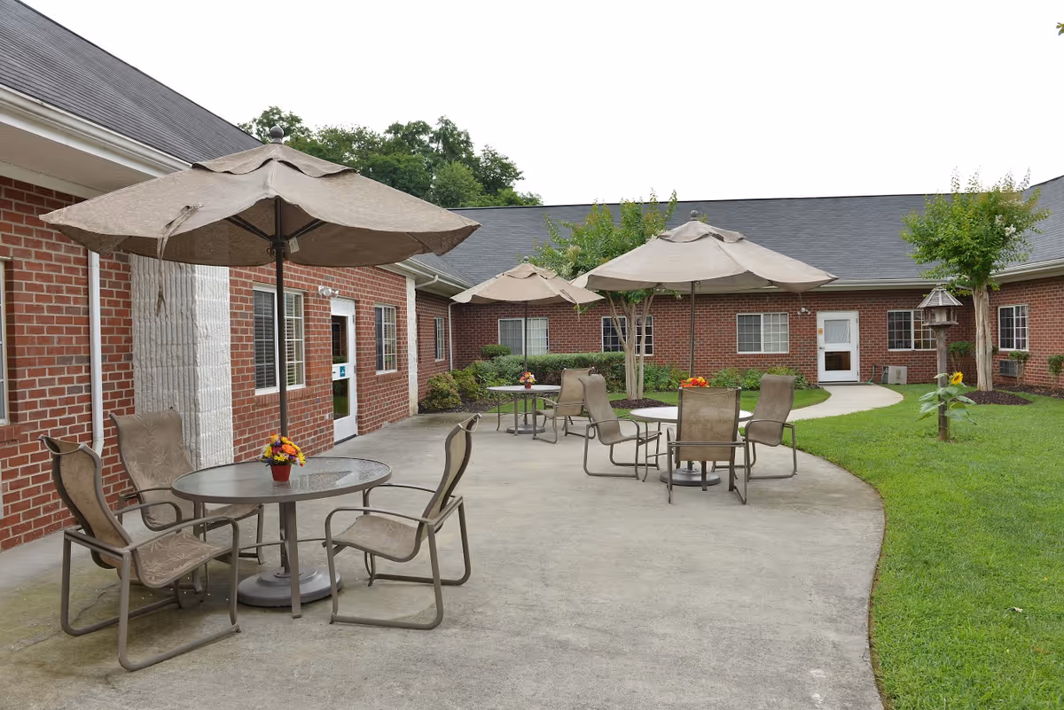 Outdoor patio area at The Laurels of Charlottesville featuring several round glass tables with beige umbrellas and matching chairs. The patio is surrounded by a red brick building with multiple windows and a door. There is a well-maintained lawn with small trees and a bird feeder on the right side.