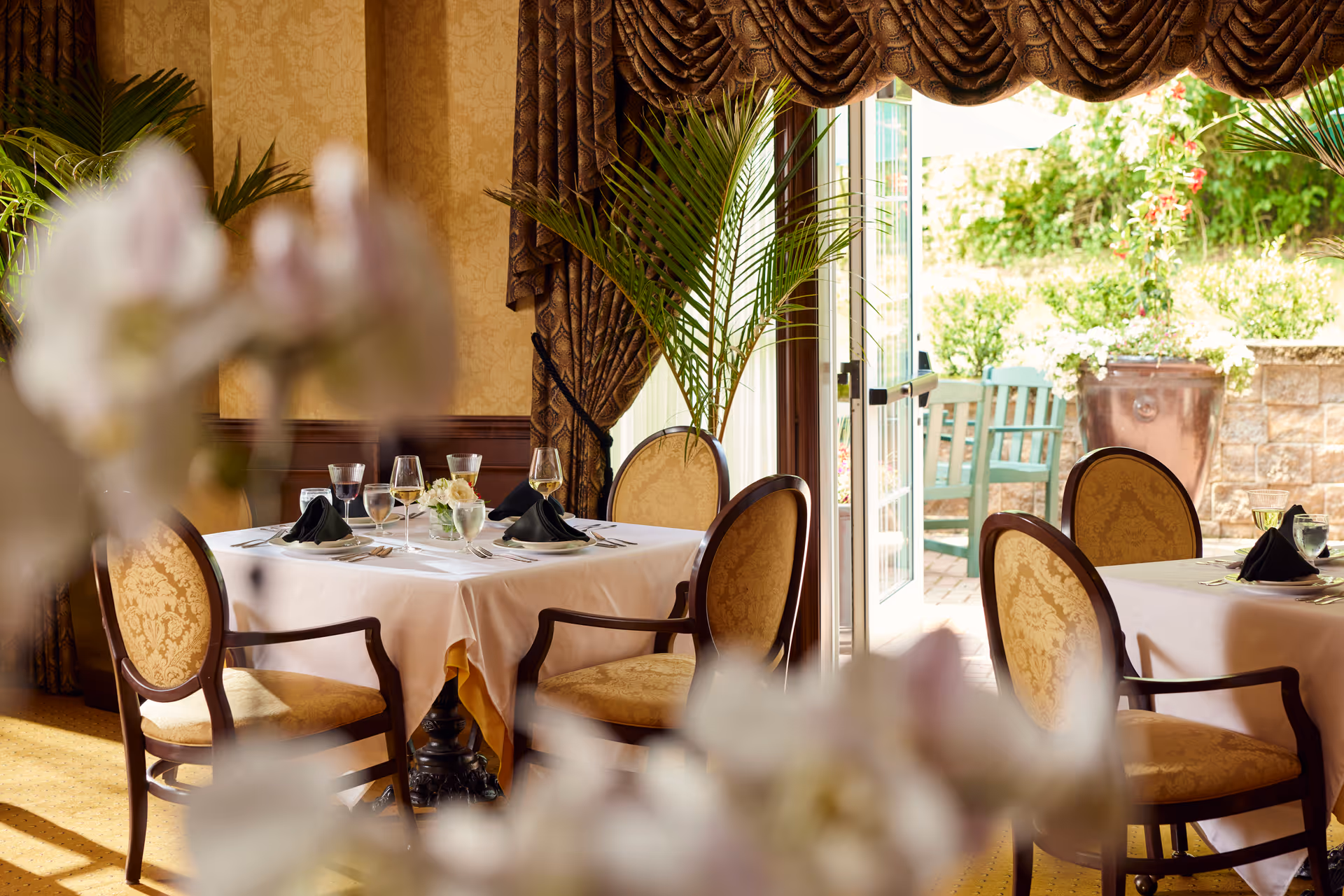 Elegant dining room with tables covered in white tablecloths, set with glassware, plates, and black napkins. The room features ornate chairs with patterned upholstery, large windows with heavy drapes, and a view of an outdoor patio with greenery and seating.