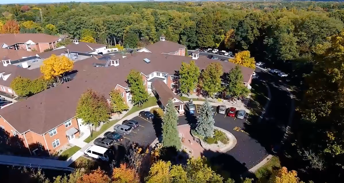 Aerial view of THE HIGHLANDS AT PITTSFORD senior living facility showing multiple connected brick buildings with brown roofs surrounded by trees with autumn foliage. There is a circular driveway with parked cars and landscaped greenery in the center. The background shows a dense forested area under a clear sky.