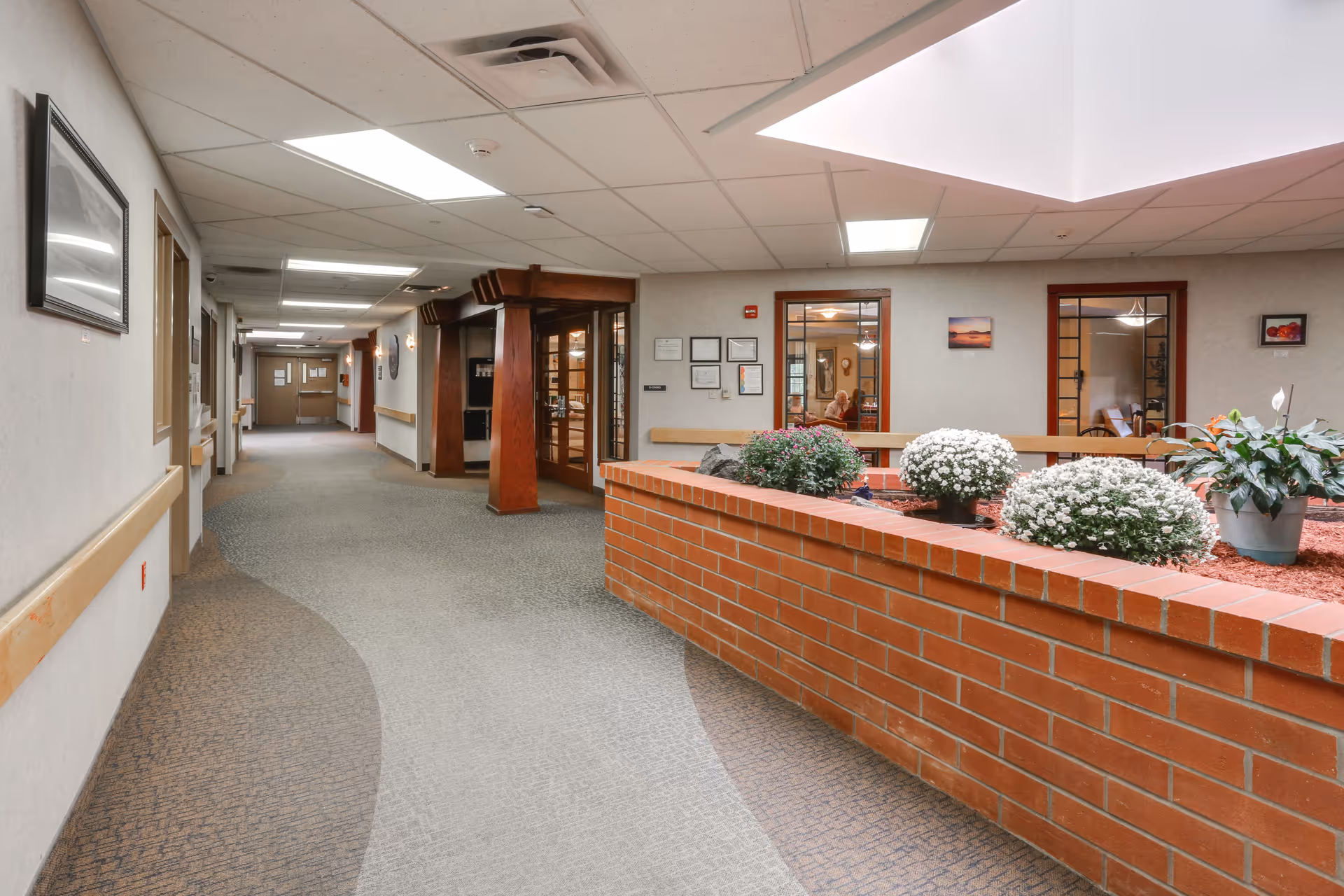 Interior hallway of a senior living facility with a carpeted floor, beige walls, and wooden handrails. A brick planter filled with green and white flowering plants is on the right side, beneath a skylight. There are framed pictures on the walls and glass doors leading to rooms in the background.