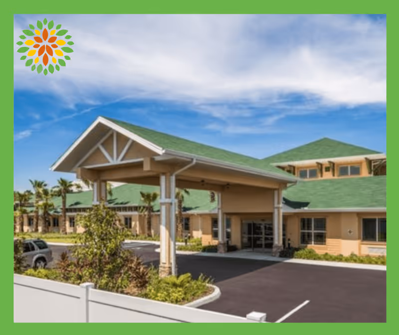 Exterior view of a senior living facility with a covered entrance, green roofs, beige walls, and surrounding landscaping including palm trees and shrubs under a partly cloudy blue sky.