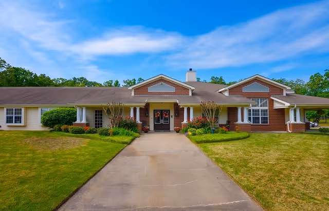 Front exterior view of a single-story senior living facility building with a brick and siding facade, a central entrance with double doors, columns, and a driveway leading up to the entrance. The building is surrounded by green lawns, shrubs, and trees under a blue sky with some clouds.