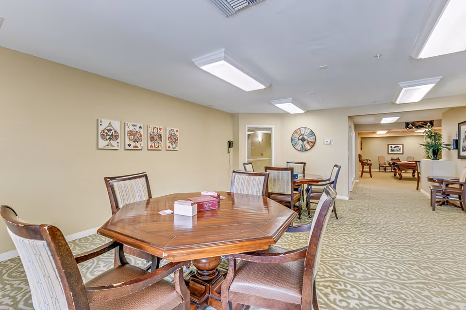 Interior view of a senior living facility common area with a wooden octagonal table surrounded by six chairs. The walls are beige with framed playing card artwork and a large decorative clock. The carpet has a patterned design, and there is a hallway leading to another room with additional seating and a pool table.