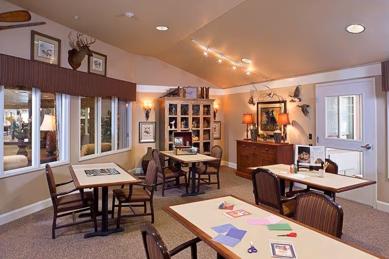 A cozy interior room with several tables and chairs arranged for activities or dining. The room features warm lighting, decorative wall-mounted animal heads, framed artwork, and a cabinet with various items. There are windows on one side and a door on the other, with craft supplies like scissors and colored paper on one table.