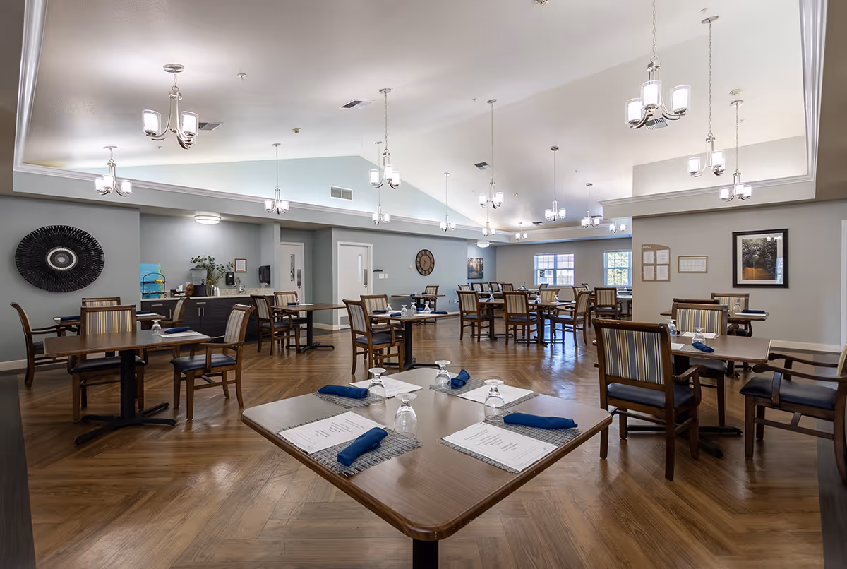 A spacious dining room in a senior living facility with multiple wooden tables and chairs arranged neatly. Each table is set with placemats, menus, upside-down glasses, and folded blue napkins. The room features a high ceiling with multiple modern chandeliers providing bright lighting. The walls are painted light gray and decorated with framed artwork and a large decorative wall piece. The floor is wooden with a herringbone pattern.