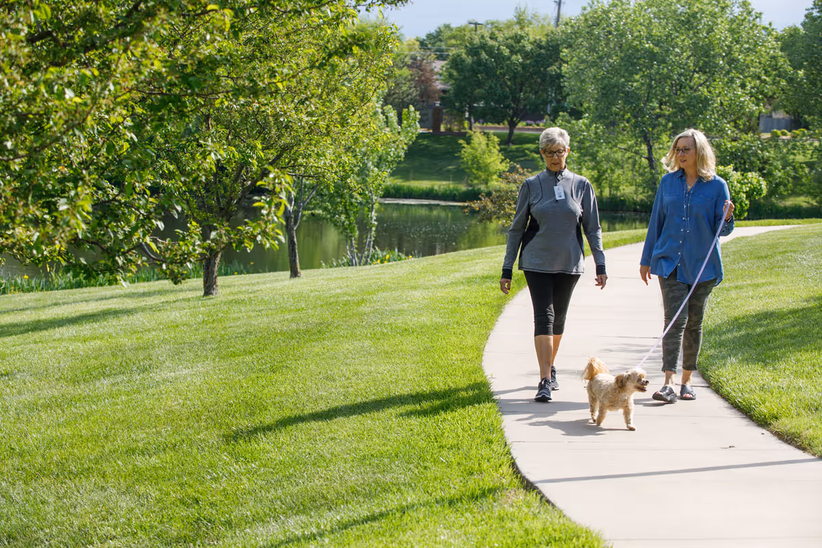 Two women walking a small dog on a leash along a paved path in a green park-like outdoor area with trees and a pond in the background.