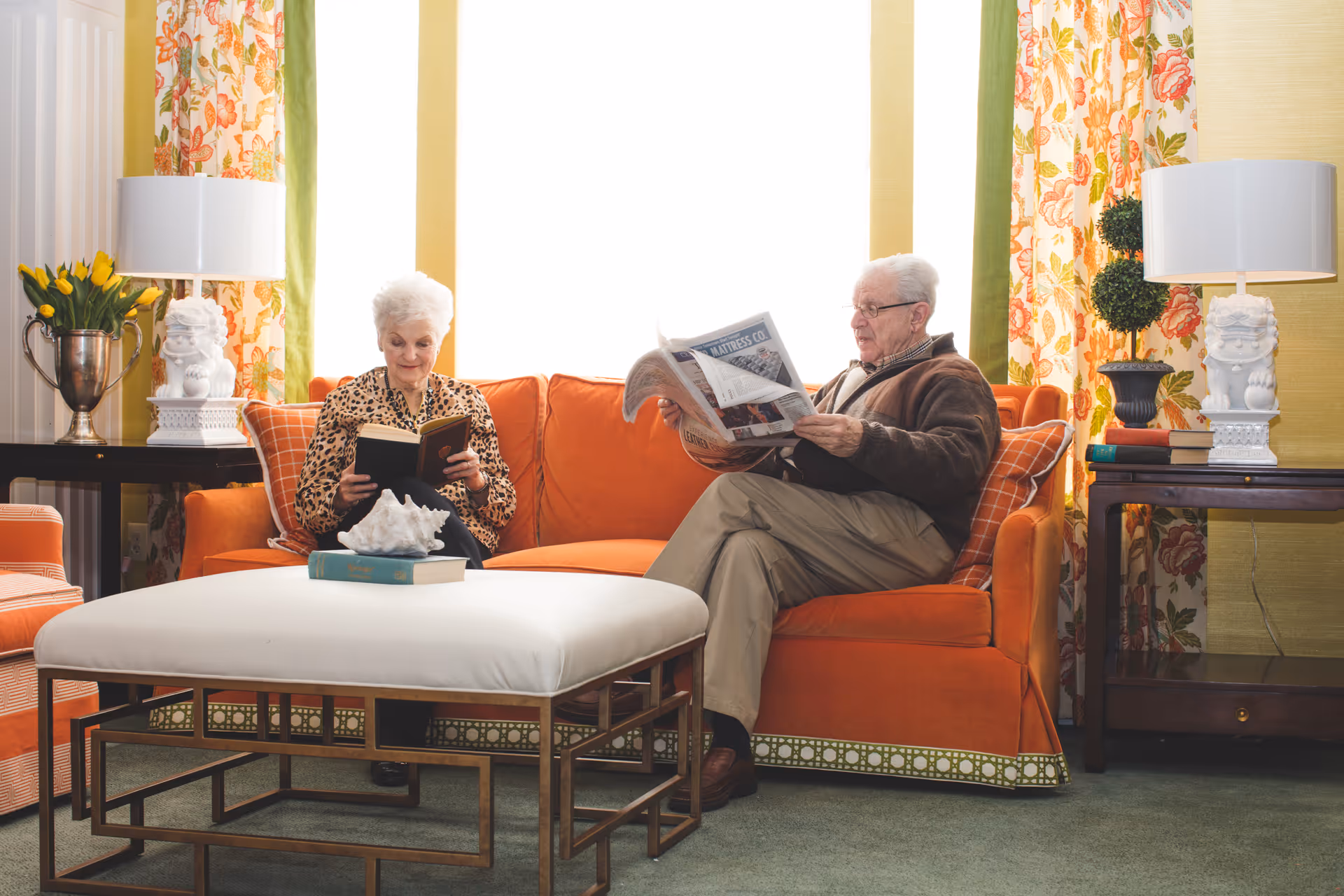 An elderly woman reads a book and an elderly man reads a newspaper while seated on an orange sofa in a cozy living room with floral curtains and a white ottoman.