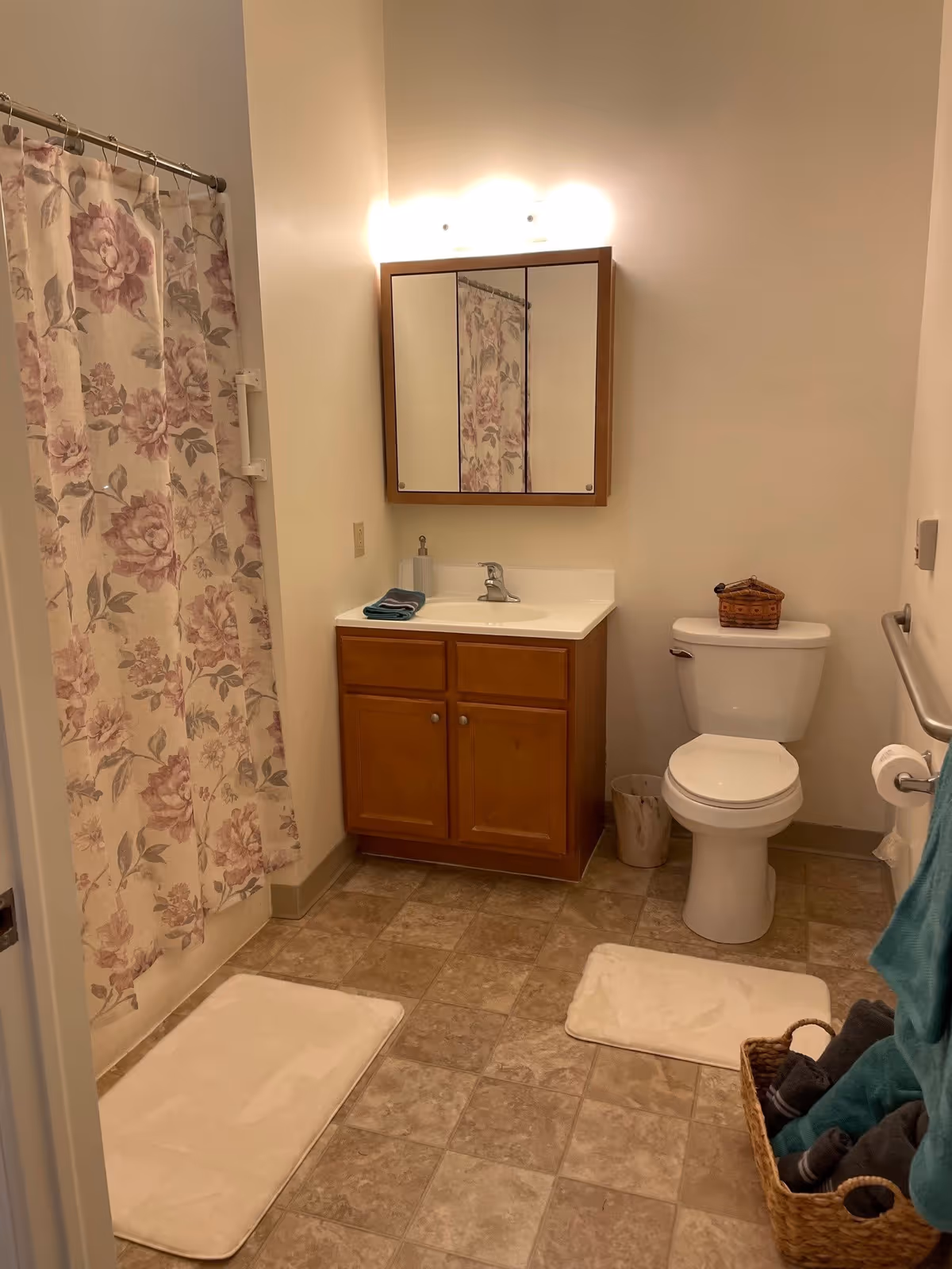 A clean bathroom with a floral shower curtain on the left, a wooden vanity with a white sink and mirror cabinet above it in the center, and a white toilet with a small basket on top on the right. There are two white bath mats on the tiled floor and a wicker basket with rolled towels next to the toilet. A towel rack with teal towels is visible on the right side.
