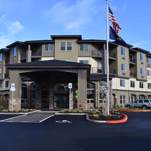 Front exterior view of a multi-story senior living facility building with a covered entrance, handicap parking spaces, and two flags on flagpoles in front under a blue sky.