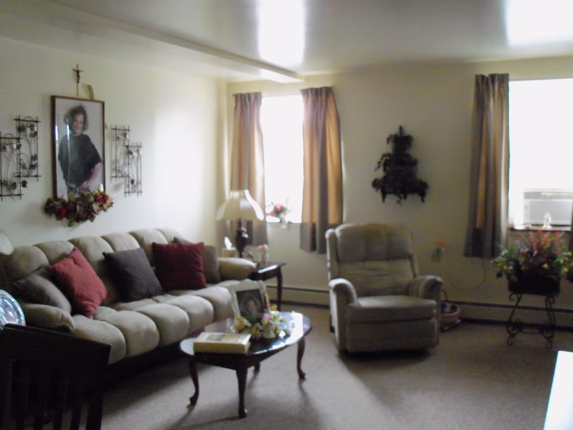 A cozy living room with a beige sofa adorned with red and brown cushions, a matching armchair, a wooden coffee table with decorative items, two windows with brown curtains, a table lamp, and wall decorations including a framed portrait and metal wall art.